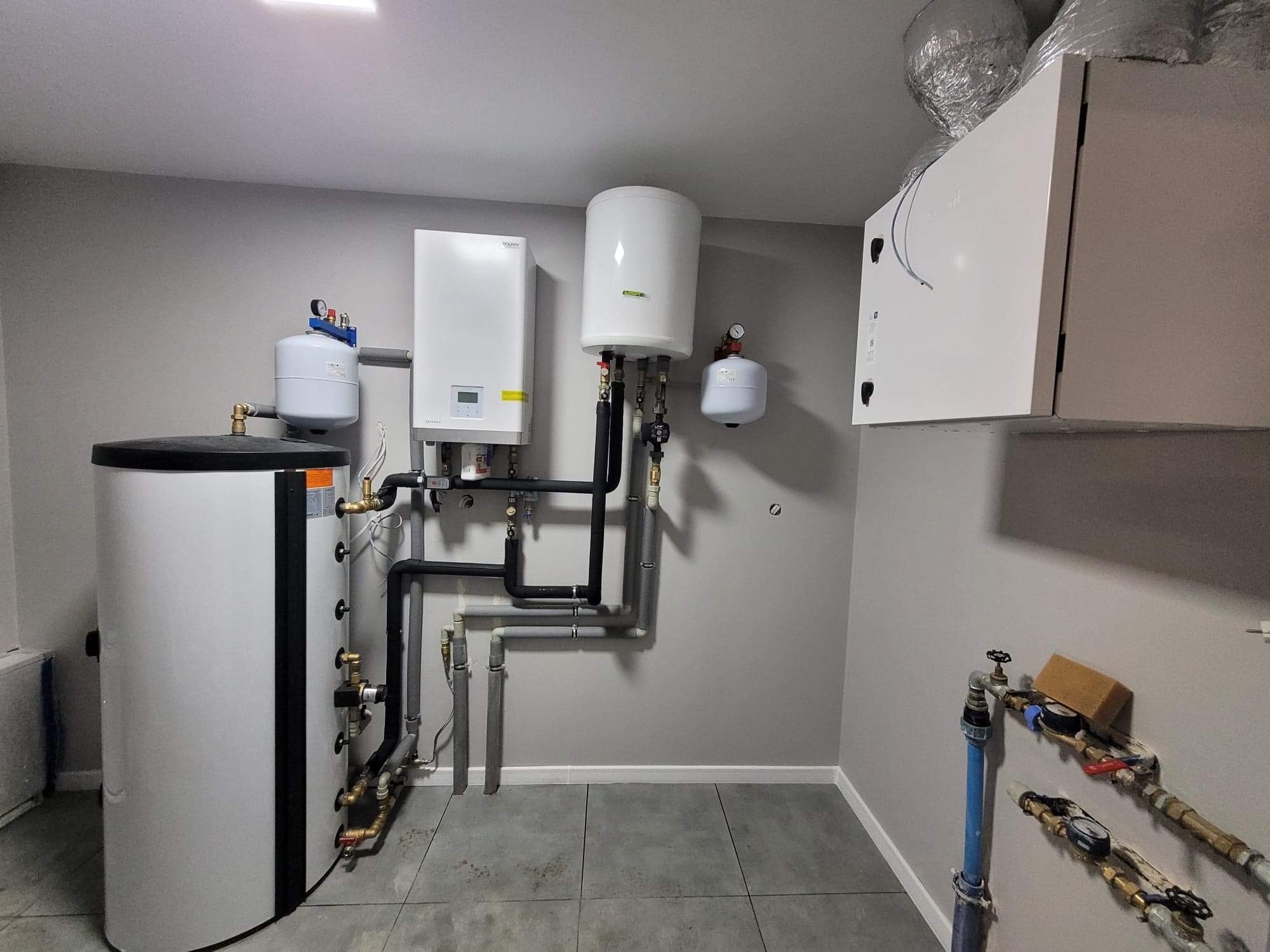 Wnętrze kotłowni z zainstalowanym zasobnikiem ciepłej wody, pompą ciepła, zbiornikiem wyrównawczym i rekuperatorem z widocznymi rurami i zaworami.