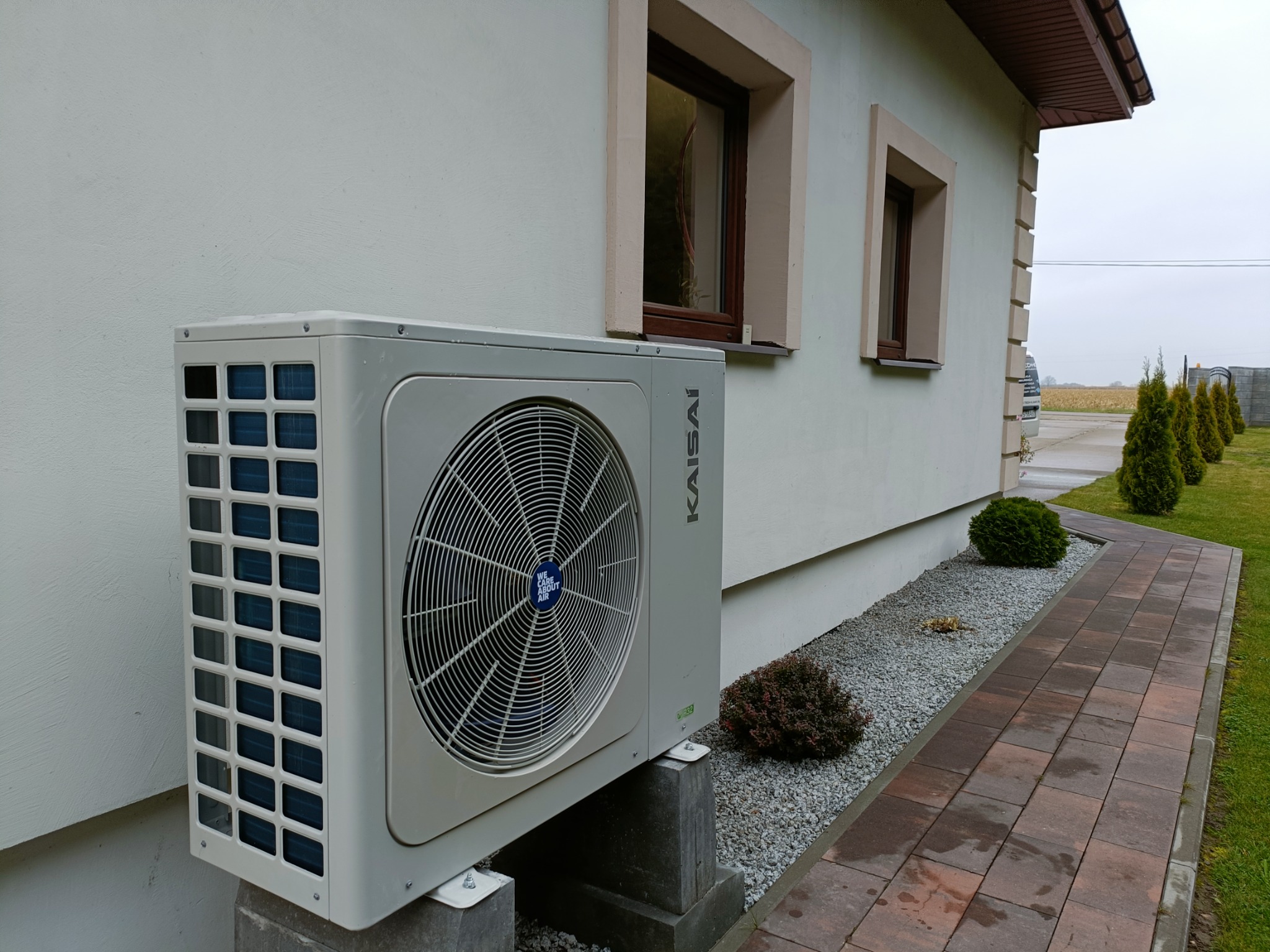 Zewnętrzna jednostka klimatyzacji Kaisai zamontowana na betonowych wspornikach przy ścianie domu z dwoma oknami, widok z boku na tle kostki brukowej i zieleni.