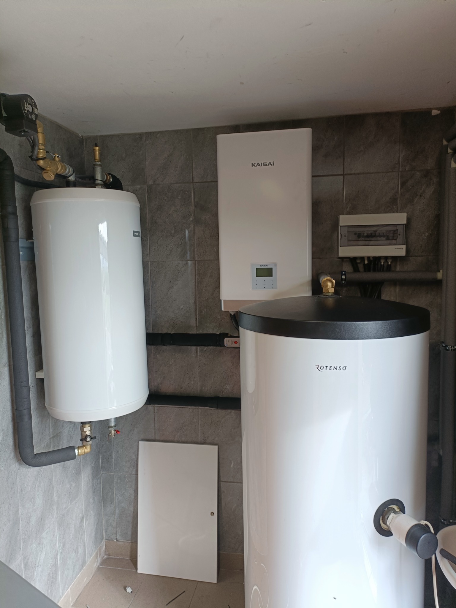 Dwa białe zbiorniki akumulacyjne Rotenso i Kaisai zamontowane na szarej, kafelkowanej ścianie w pomieszczeniu technicznym, rury izolowane termicznie.