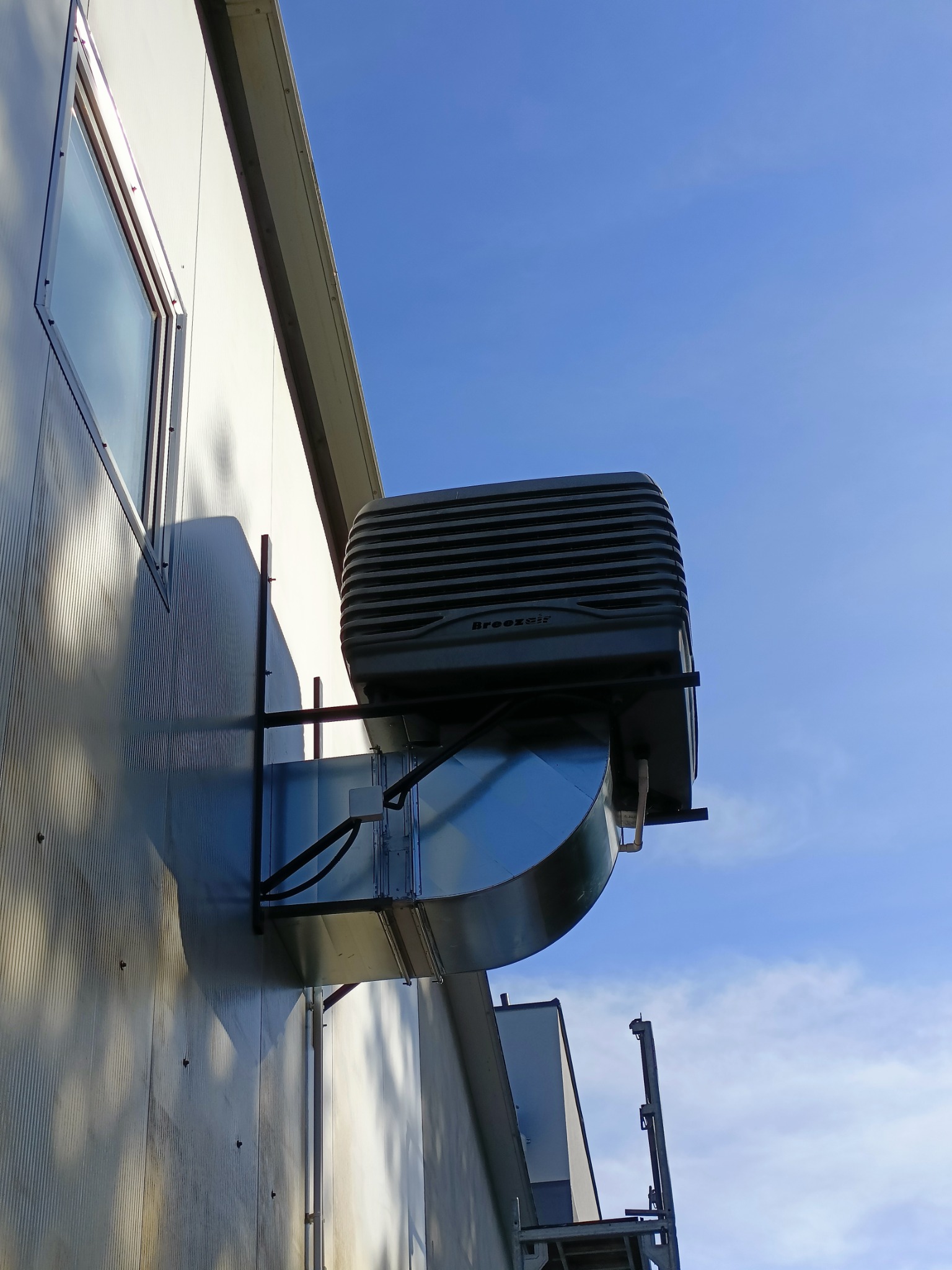 Szara jednostka rekuperacji Breezair zamontowana na zewnątrz budynku z blachy trapezowej, widoczny kanał wentylacyjny i fragment okna na tle błękitnego nieba.