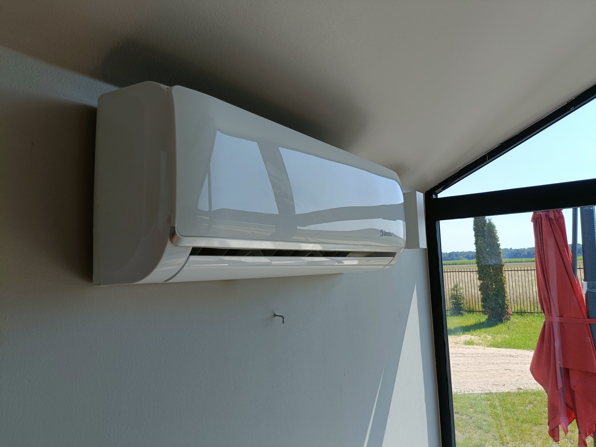 Biały klimatyzator ścienny marki Beretta zamontowany w pomieszczeniu z widokiem na zewnątrz przez duże okno; hak w ścianie pod urządzeniem.