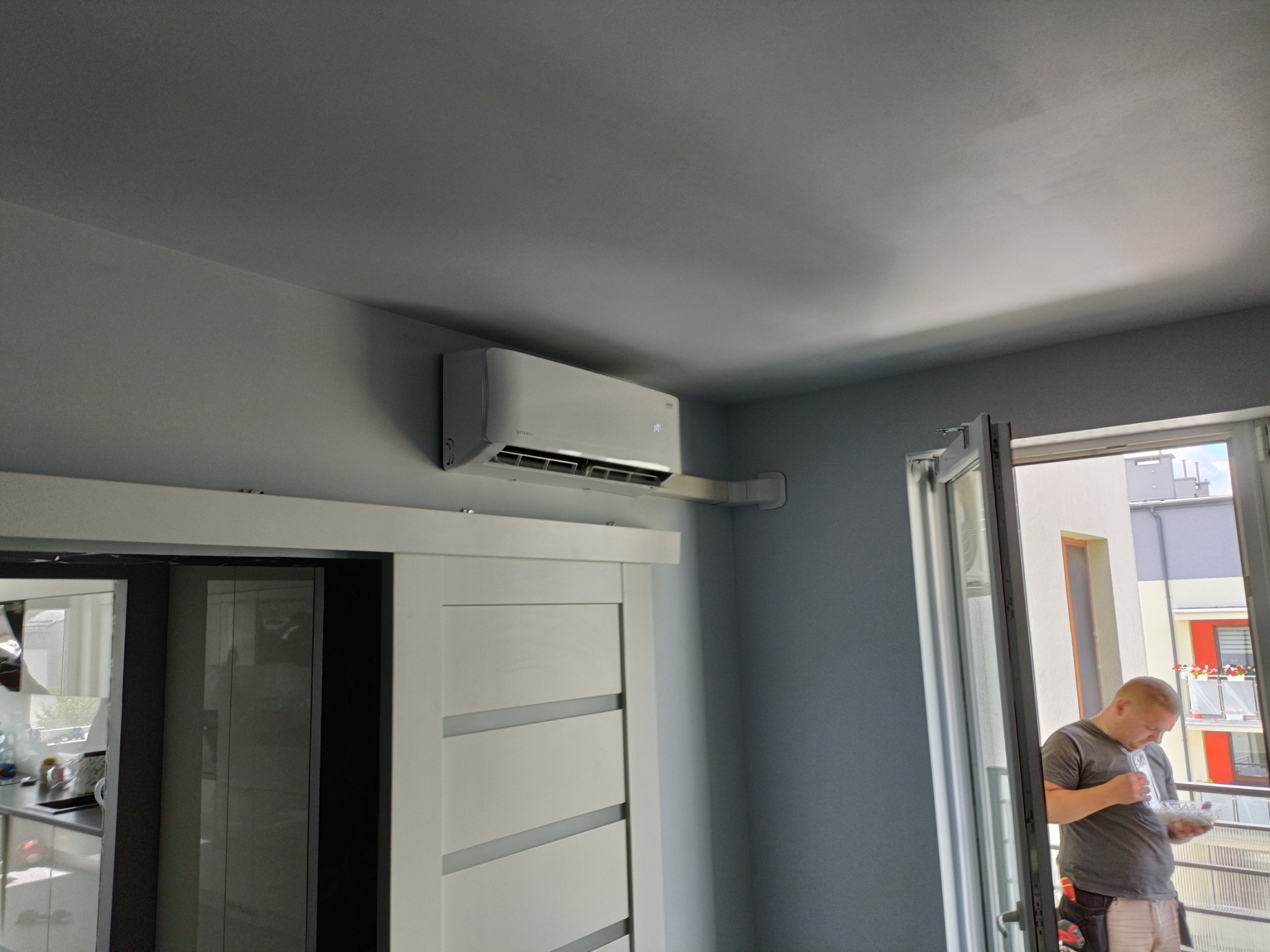 Nowoczesny klimatyzator ścienny w szarym pomieszczeniu, widoczny pracownik na balkonie w tle, spożywający posiłek.