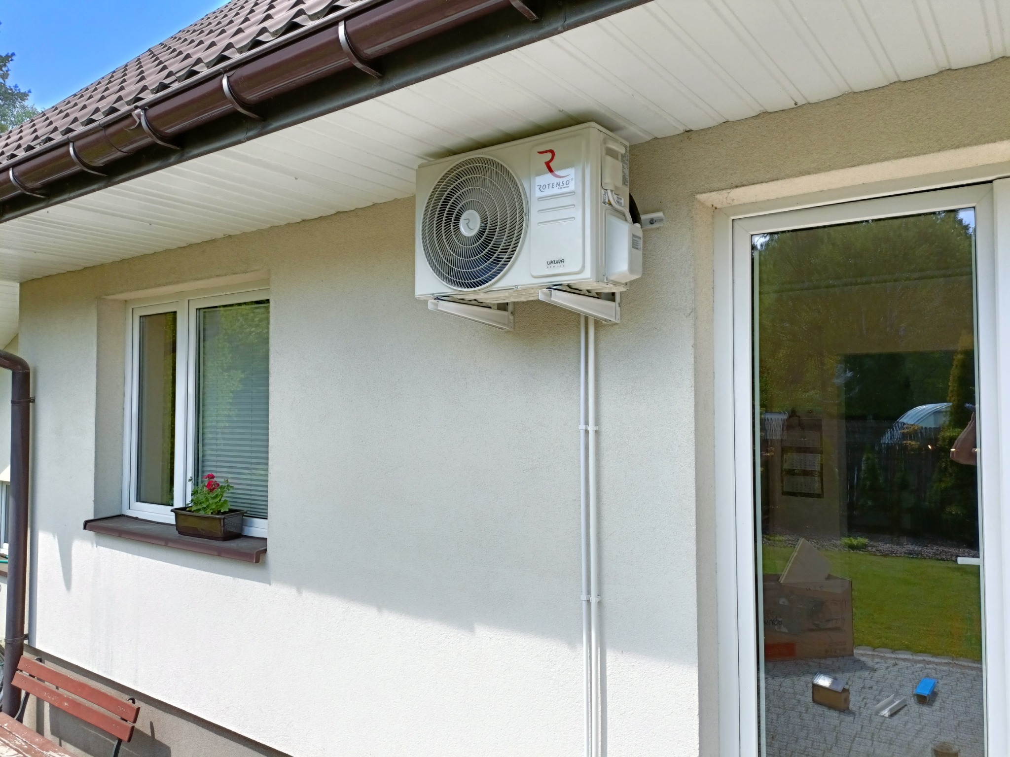 Zewnętrzna jednostka klimatyzacji Rotenso Ukura zamontowana na jasnej ścianie domu, widoczne rury prowadzące w dół, okno z kwiatami i odbicie ogrodu w szybie drzwi balkonowych.