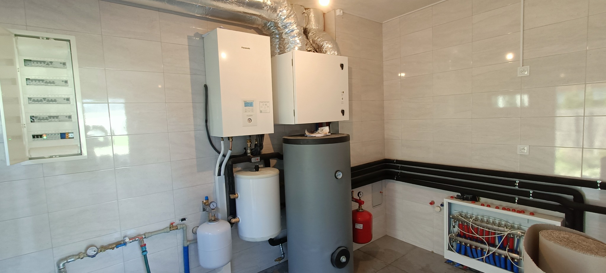 Zestaw urządzeń grzewczych w kotłowni z białymi płytkami na ścianach, widoczny biały piec Panasonic, szary zasobnik ciepłej wody, czerwona butla wyrównawcza i rozdzielacz podłogówki...