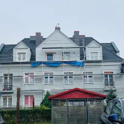 Wielopiętrowy budynek w trakcie prac elewacyjnych, otoczony rusztowaniem, z widocznymi niebieskimi plandekami zabezpieczającymi okna, pracownik na rusztowaniu, w tle zaparkowane samochody i zieleń.