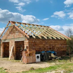 Budowa domu z cegły ceramicznej: ściany wzniesione do poziomu dachu, widoczna konstrukcja więźby dachowej, na pierwszym planie stos cegieł i pralka.