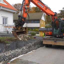 ADDO Sp. z o.o. - Pomarańczowa koparka Doosan umieszcza kamienie w metalowej siatce gabionowej, tworząc mur oporowy na posesji z białym domem w tle, pracownik w odblaskowej kamizelce nadzoruje proces.