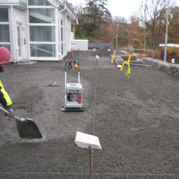 ADDO Sp. z o.o. - Pracownik w odzieży roboczej wyrównuje podłoże z drobnego kruszywa łopatą, w tle zagęszczarka wibracyjna, wytyczone sznurki i ogrodzenie gabionowe z żółtymi rurami ochronnymi.