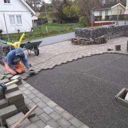ADDO Sp. z o.o. - Pracownik w pomarańczowych spodniach i niebieskiej bluzie układa szare kostki brukowe na przygotowanym podłożu z drobnego żwiru, obok leżą słuchawki ochronne i poziomica, w tle widoczny dom...