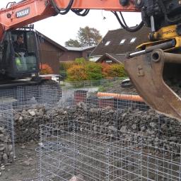 ADDO Sp. z o.o. - Pomarańczowa koparka Doosan z operatorem wypełnia kamieniami metalowe kosze gabionowe na placu budowy, w tle budynki mieszkalne i krzewy.