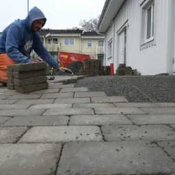 Pracownik w niebieskiej bluzie z kapturem i pomarańczowych spodniach klęczy układając szare kostki brukowe na przygotowanym podłożu z drobnego żwiru, w tle budynki mieszkalne i kontener na gruz.