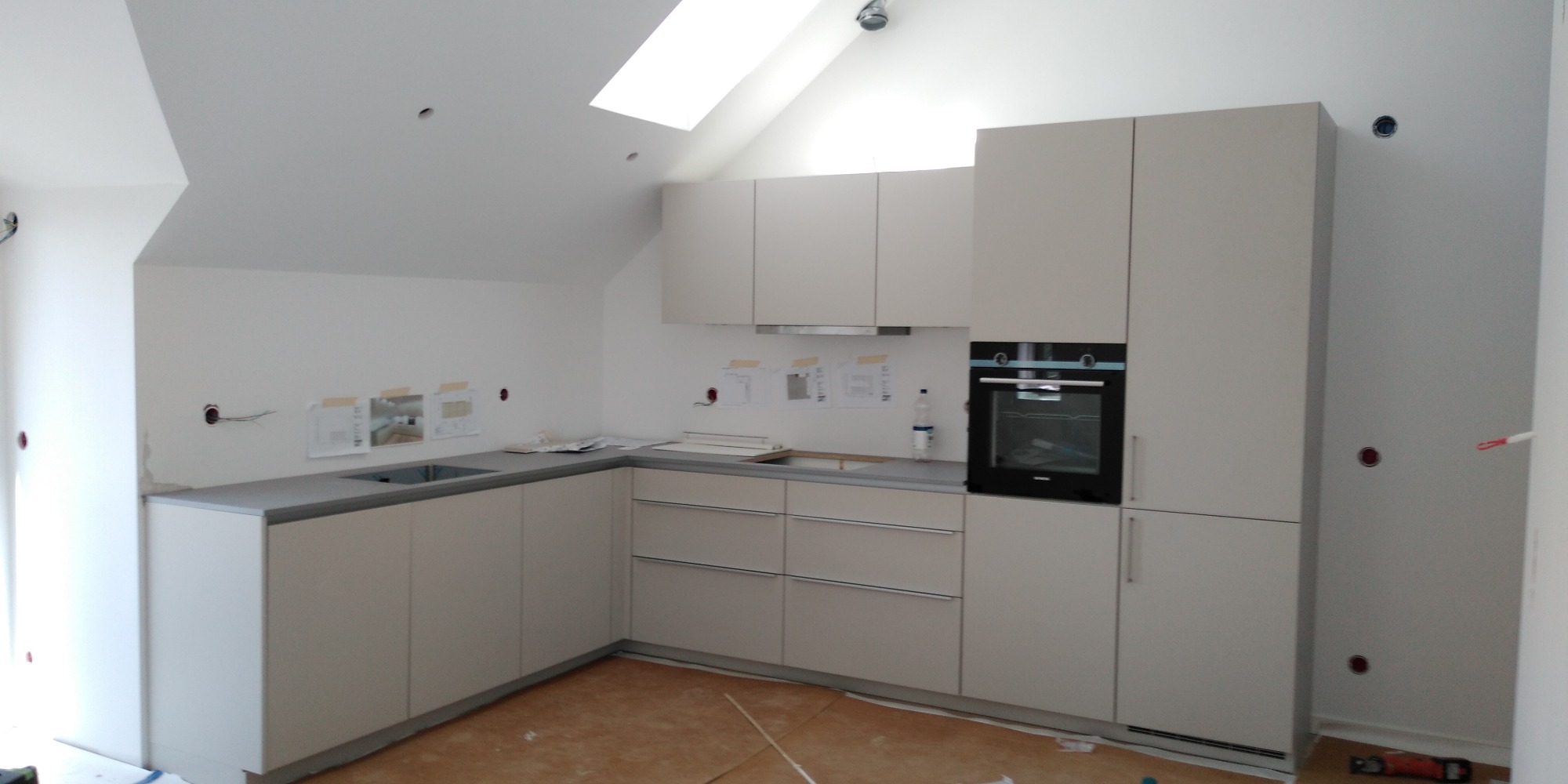 Nowa, jasna kuchnia w trakcie montażu, widoczne przygotowania do podłączenia instalacji elektrycznej i sanitarnej, kartki z planami na ścianie.