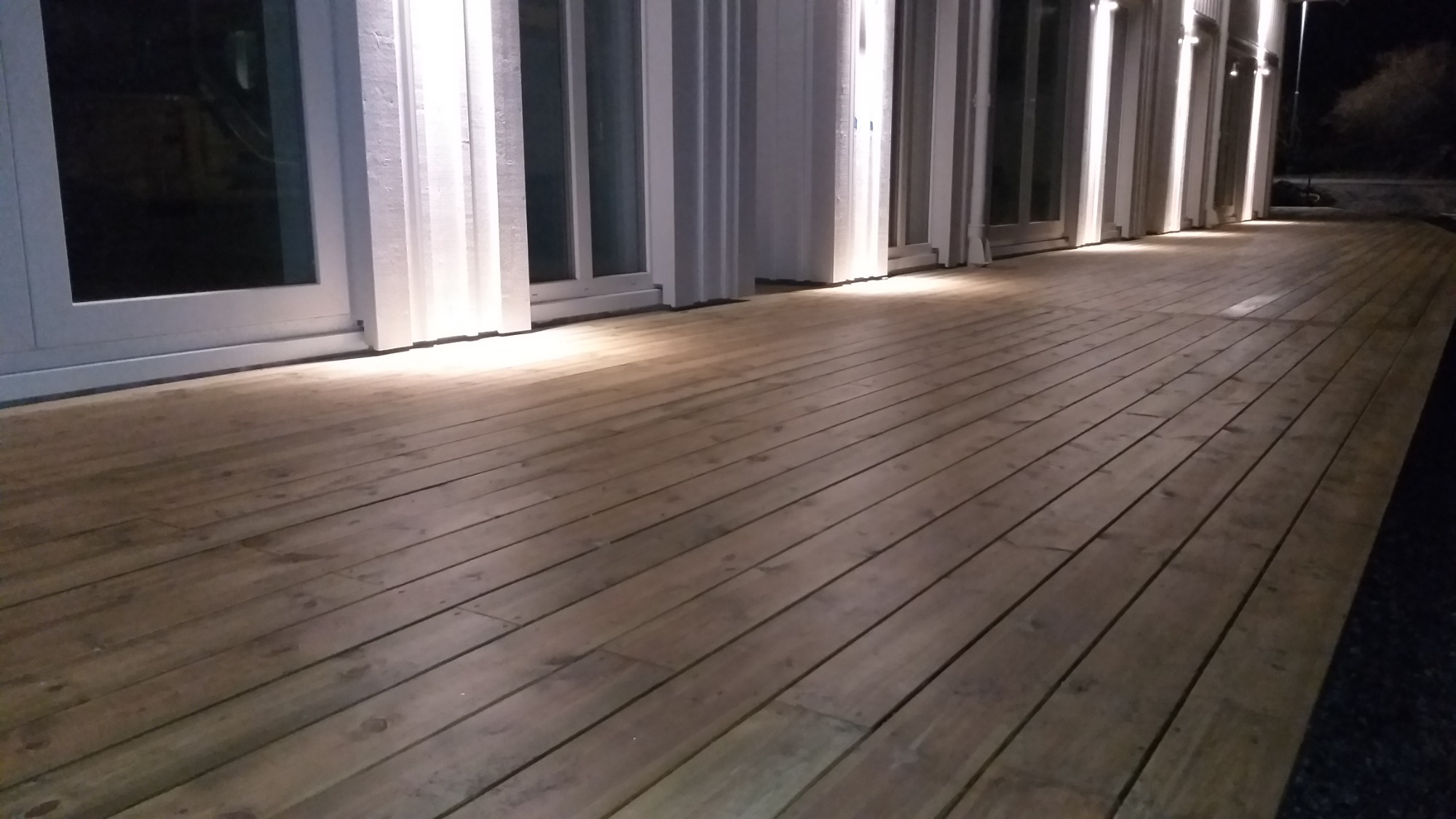 Drewniany taras oświetlony punktowymi lampami elewacyjnymi, widoczne białe ramy okien i drzwi balkonowych w tle nocnego nieba.