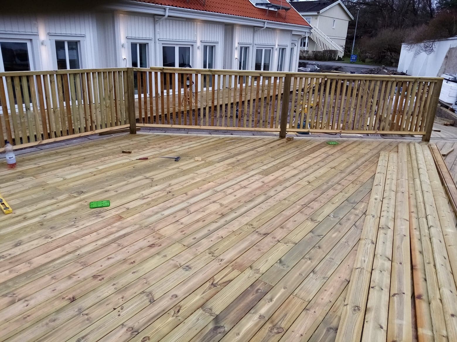 Budowa drewnianego tarasu z balustradą przy jasnym domu jednorodzinnym, widoczne narzędzia i butelka wody.