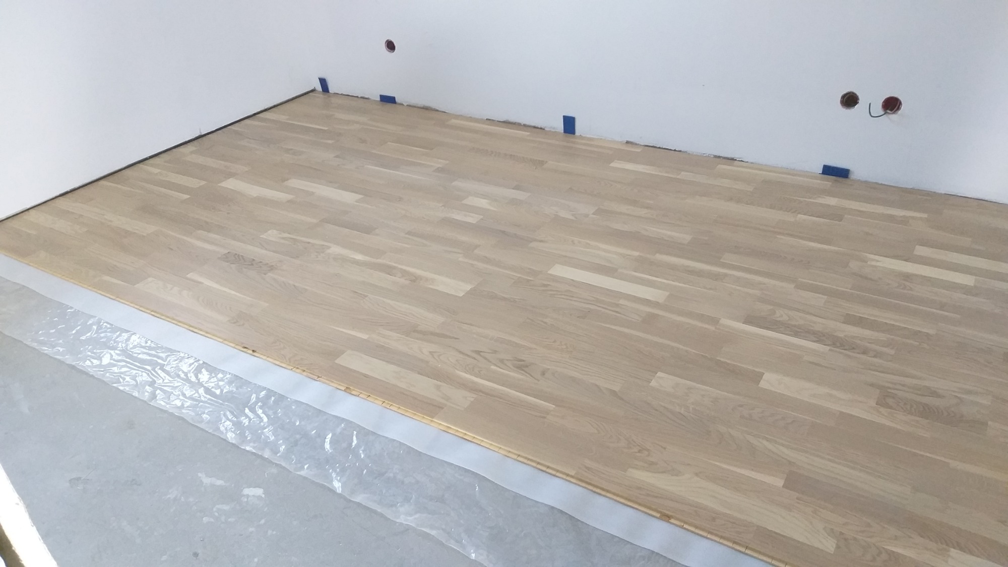 Układanie jasnego parkietu w pokoju, widoczne zabezpieczenie folią podłoża, nieukończona podłoga, przygotowane gniazdka elektryczne w ścianie.