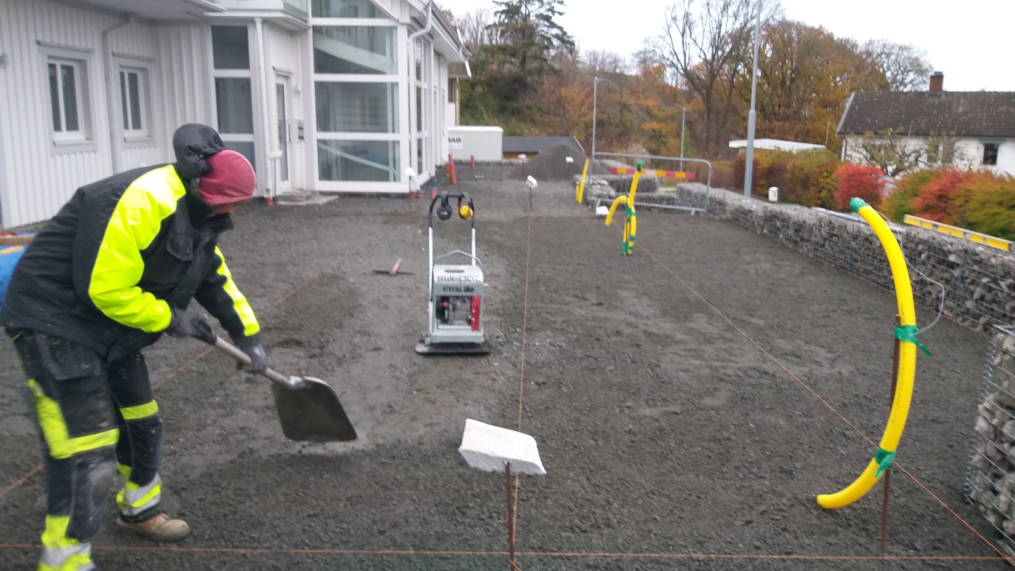 Pracownik w odzieży roboczej wyrównuje podłoże z drobnego kruszywa łopatą, w tle zagęszczarka wibracyjna, wytyczone sznurki i ogrodzenie gabionowe z żółtymi rurami ochronnymi.