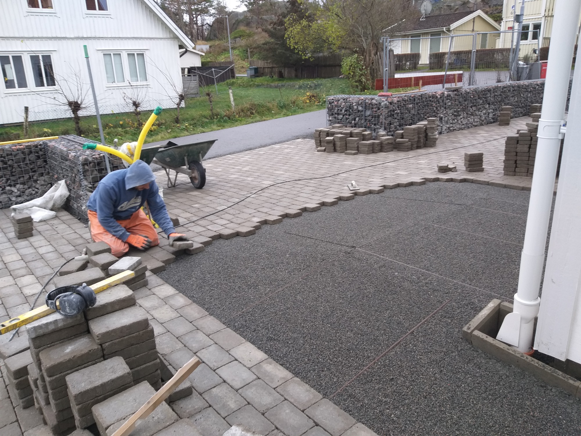 Pracownik w pomarańczowych spodniach i niebieskiej bluzie układa szare kostki brukowe na przygotowanym podłożu z drobnego żwiru, obok leżą słuchawki ochronne i poziomica, w tle widoczny dom...