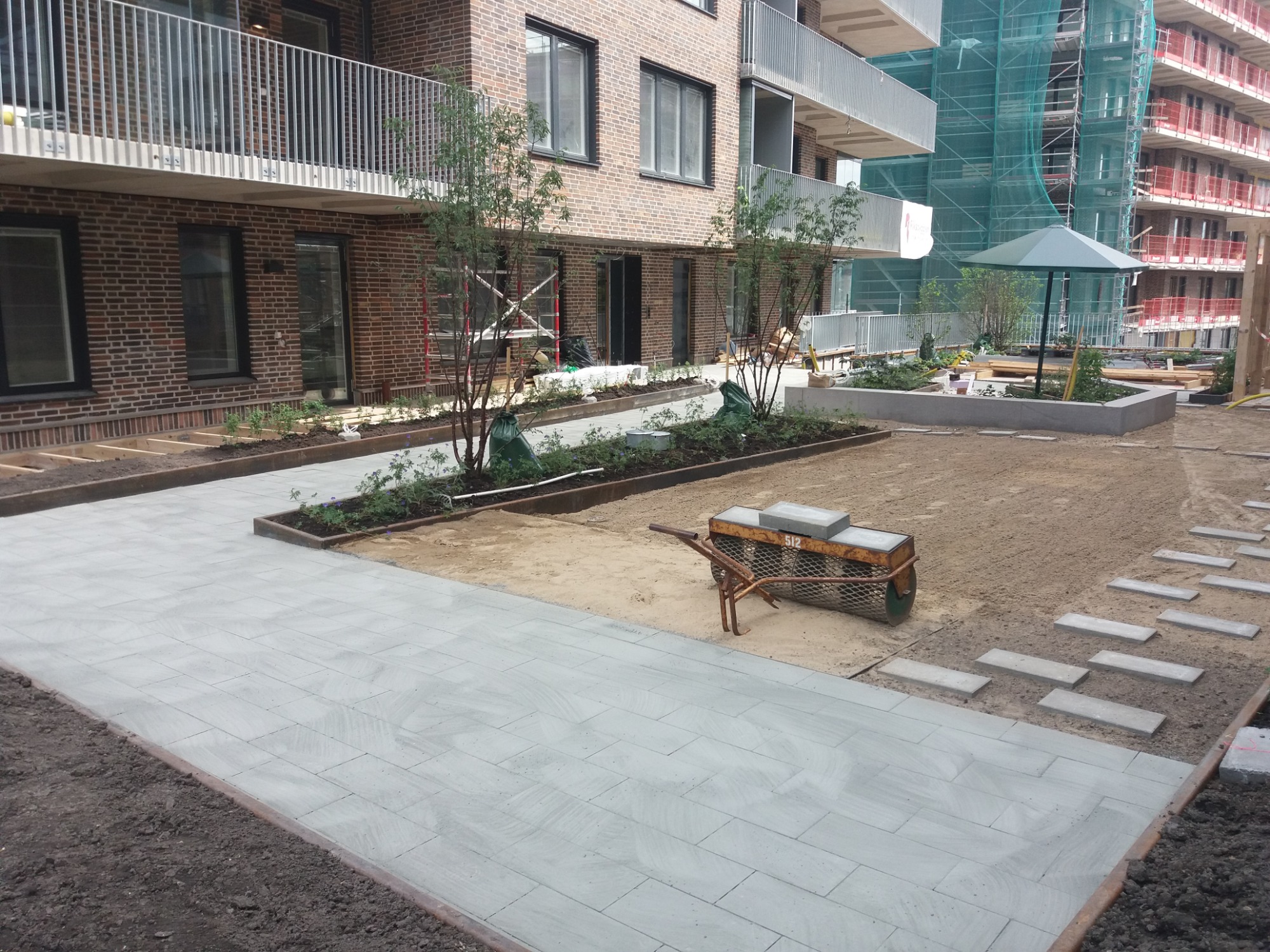 Świeżo ułożona, jasnoszara kostka brukowa prowadzi przez teren budowy nowego osiedla; widoczna taczka z kostką, rabaty z sadzonkami i budynek w rusztowaniach.