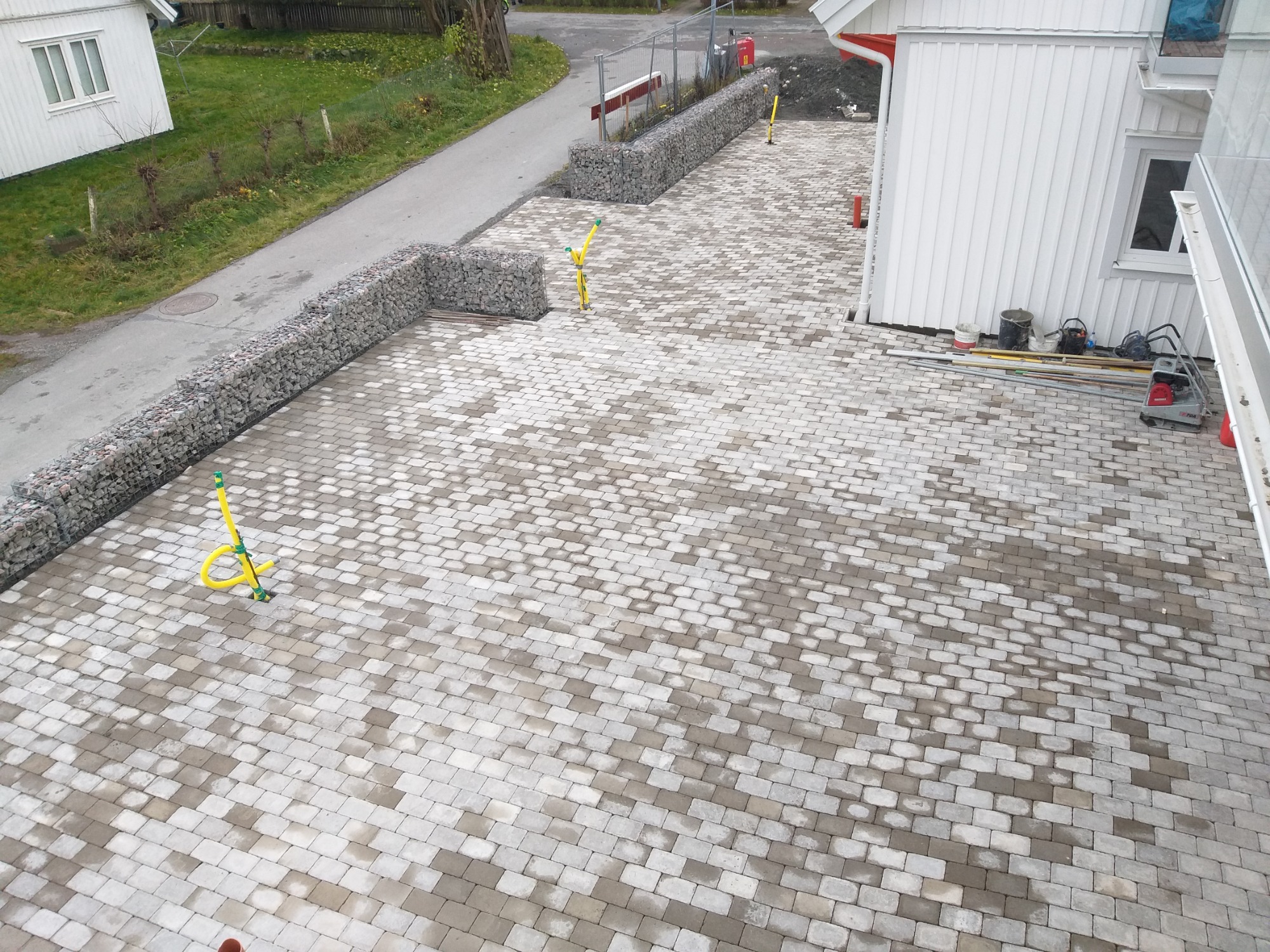 Świeżo ułożona kostka brukowa w odcieniach szarości, z widocznymi żółtymi rurami instalacji i gabionami z kamienia w tle, częściowo mokra powierzchnia kostki.