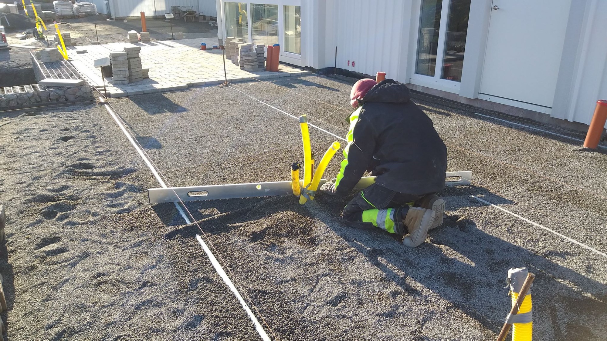 Pracownik w odzieży roboczej klęczy na podsypce z kruszywa, wyrównując powierzchnię łatą aluminiową, widoczne sznurki wyznaczające poziom i ułożone obrzeża z kostki.