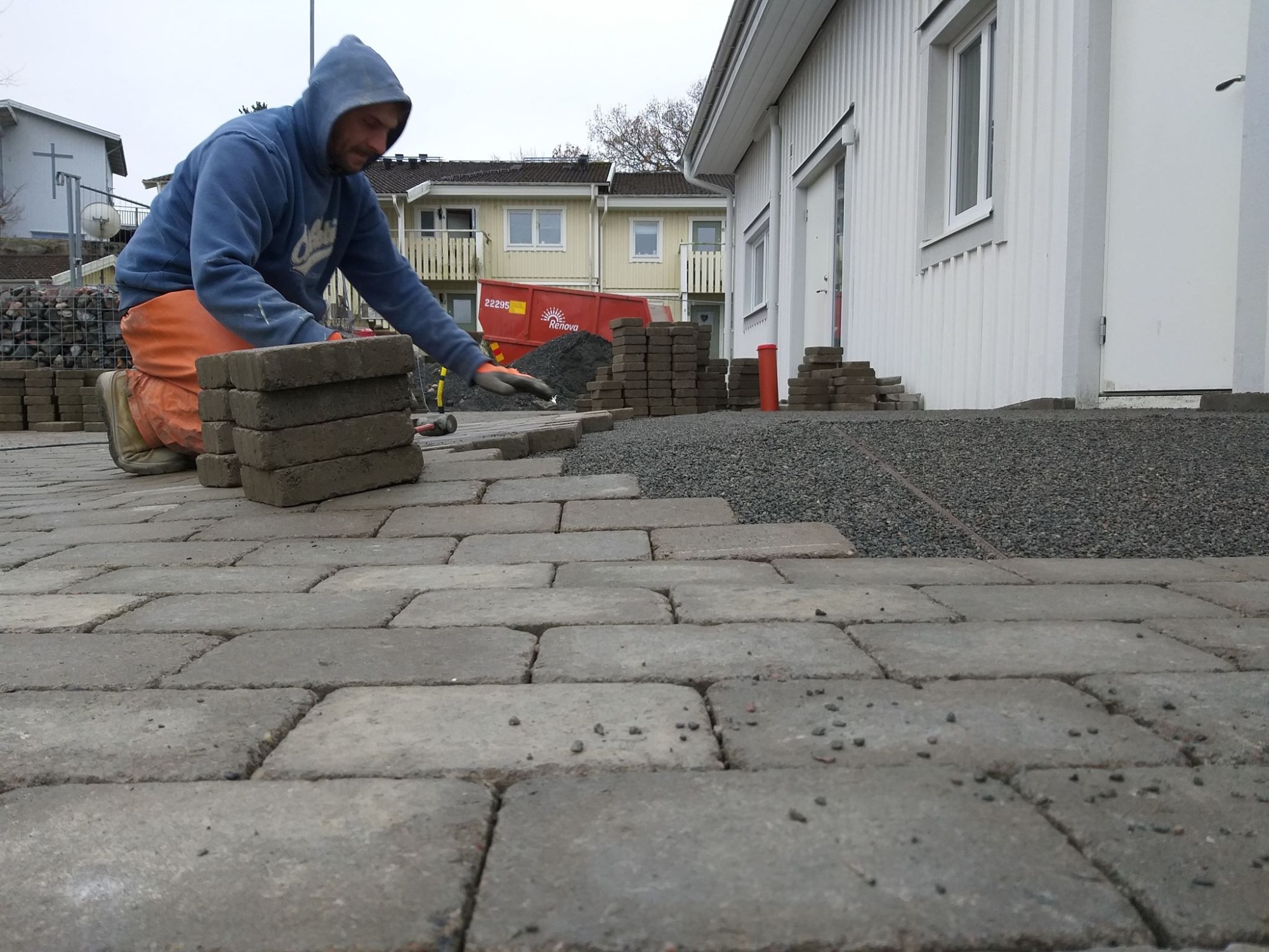 Pracownik w niebieskiej bluzie z kapturem i pomarańczowych spodniach klęczy układając szare kostki brukowe na przygotowanym podłożu z drobnego żwiru, w tle budynki mieszkalne i kontener na gruz.