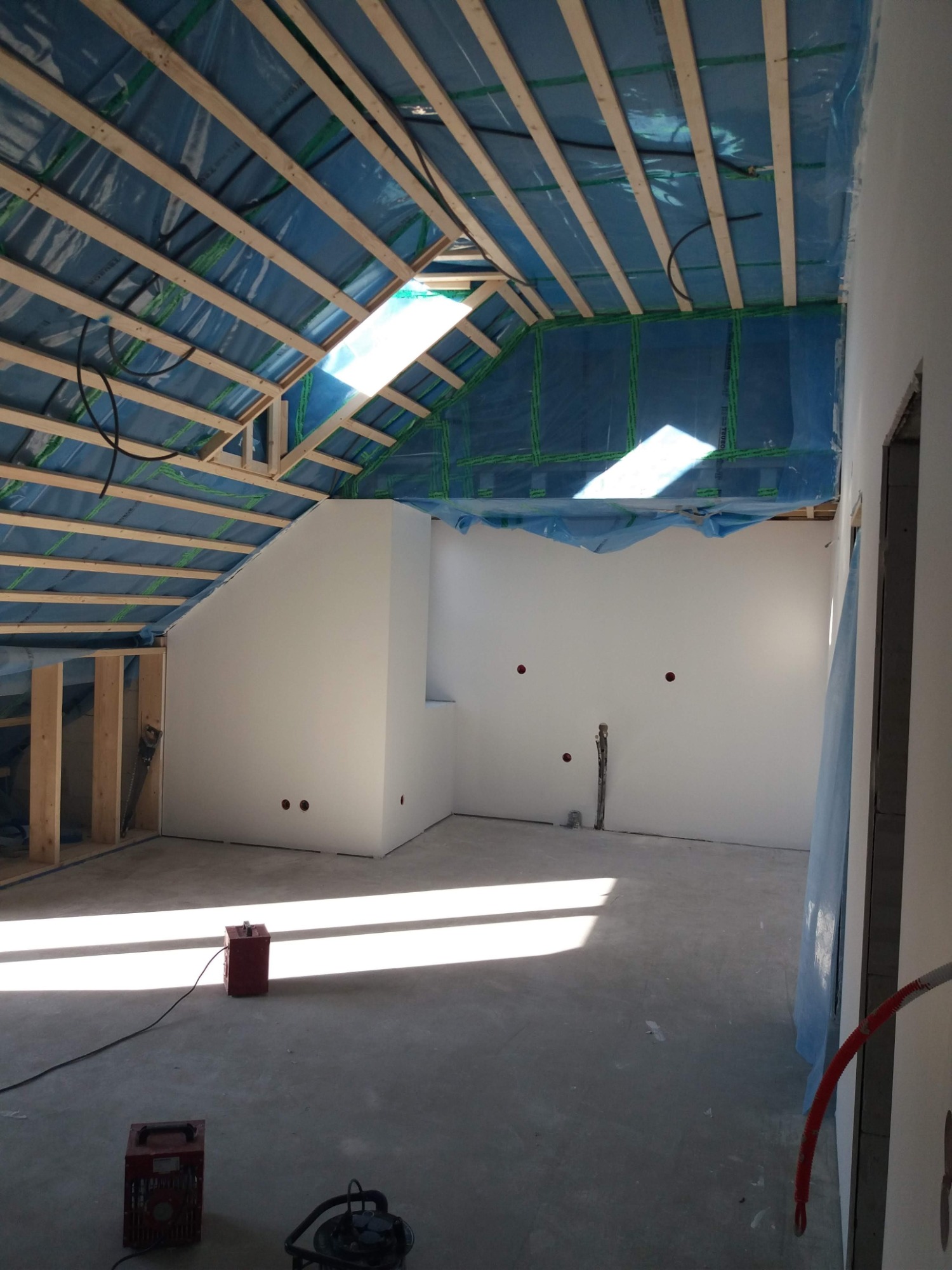 Wnętrze poddasza w trakcie prac wykończeniowych w Warszawie, widoczne drewniane elementy konstrukcyjne dachu zabezpieczone folią, gładkie, białe ściany oraz sprzęt budowlany na podłodze.