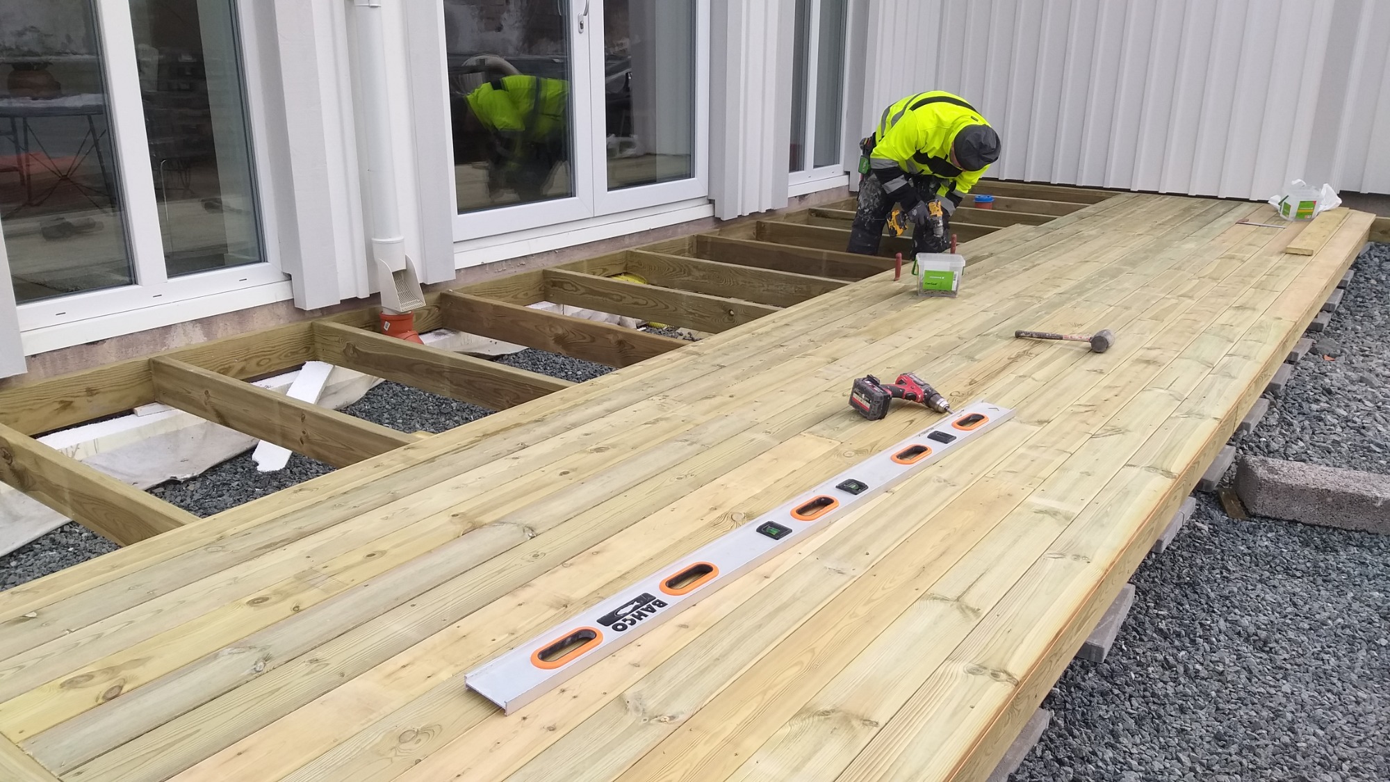 Pracownik w odblaskowej kurtce montuje drewniany taras przy białym budynku. Widoczne narzędzia: poziomica Bahco, wkrętarka, młotek. Część konstrukcji tarasu jeszcze nieukończona.