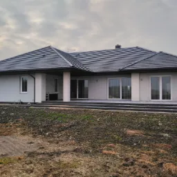 Elewacja parterowego domu jednorodzinnego z ciemną dach&oacute;wką, widok z zewnątrz, w tle pochmurne niebo.