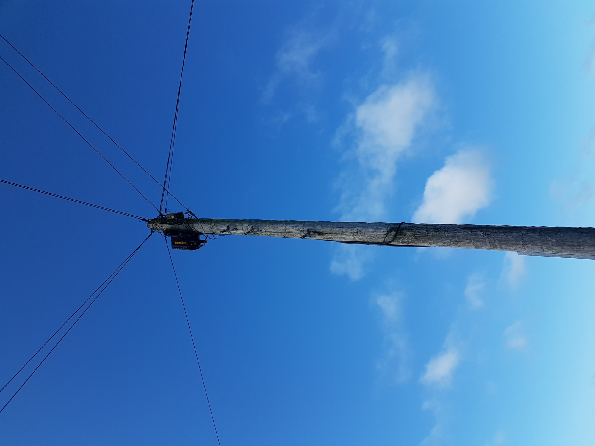 Słup telekomunikacyjny widziany z dołu na tle błękitnego nieba z delikatnymi obłokami, widoczne przewody i zamontowane urządzenia.