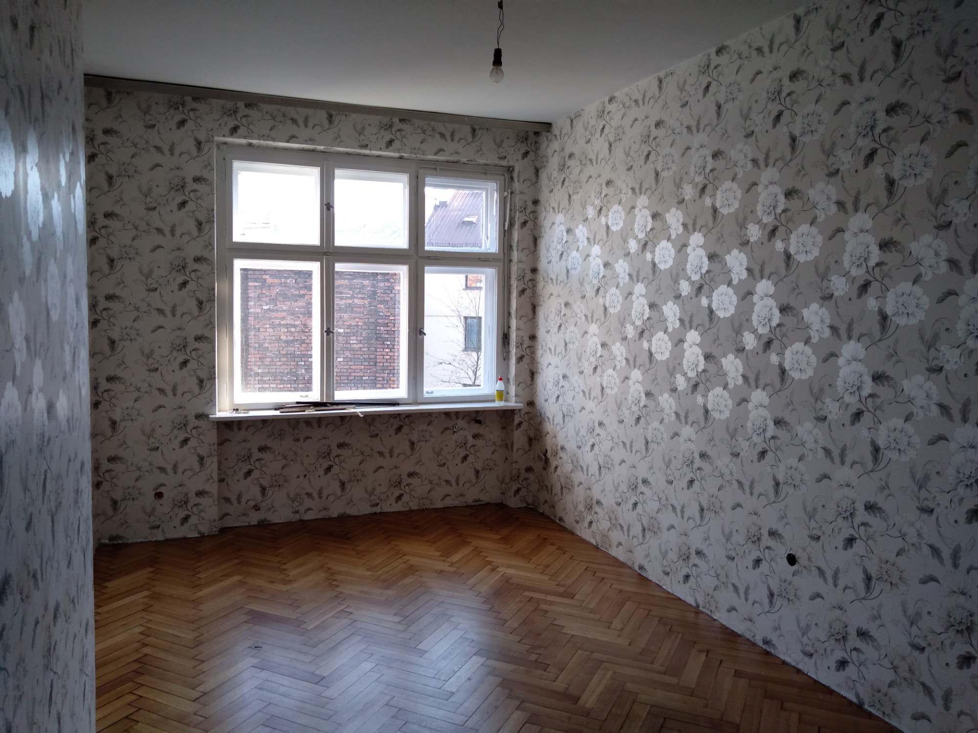 Pusty pokój po tapetowaniu w kwiaty, parkiet w jodełkę, okno z widokiem na ceglany budynek, surowa żarówka zwisająca z sufitu.