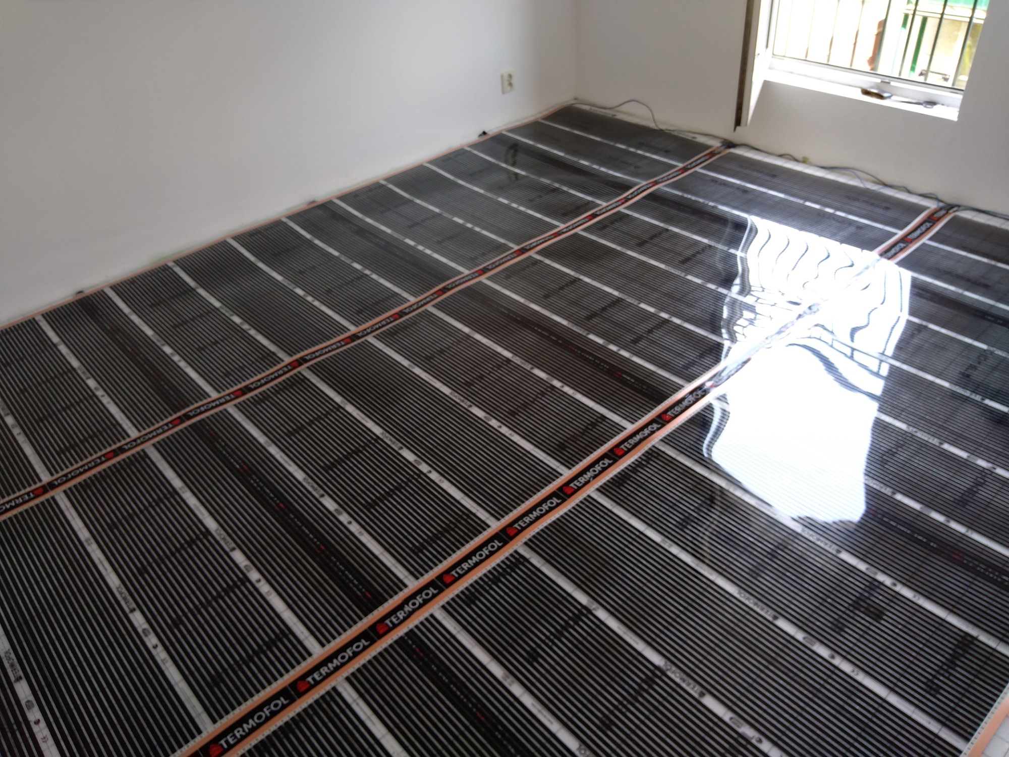 Rozłożona mata grzewcza Termofol na podłodze w jasnym pomieszczeniu, przygotowana do montażu ogrzewania podłogowego, widoczne kable i gniazdko elektryczne w tle.