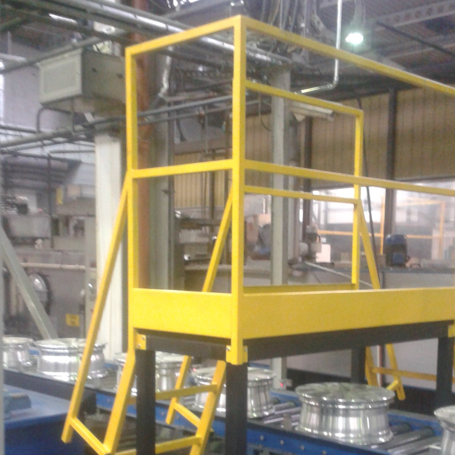Żółta, metalowa konstrukcja pomostu roboczego z drabinką w hali produkcyjnej z elementami linii produkcyjnej w tle.