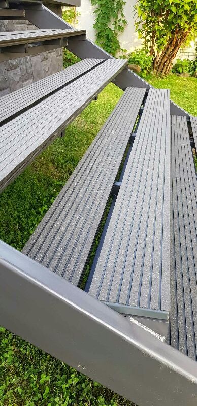 Metalowe schody zewnętrzne z szarymi ryflowanymi stopniami, widok z dołu na tle zielonej trawy i roślinności.