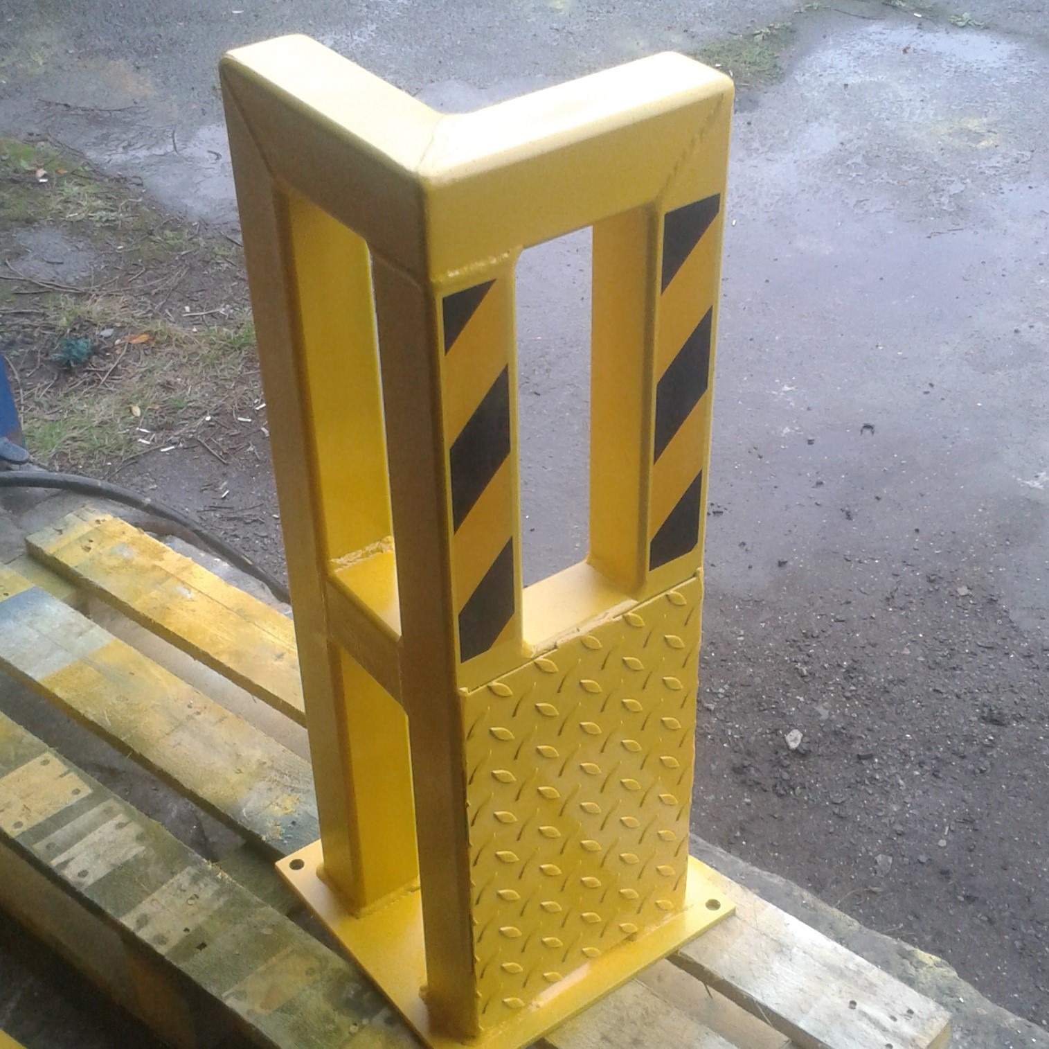 Żółty, stalowy odbojnik narożny z czarnymi pasami ostrzegawczymi, przymocowany do drewnianej palety, widok z bliska.