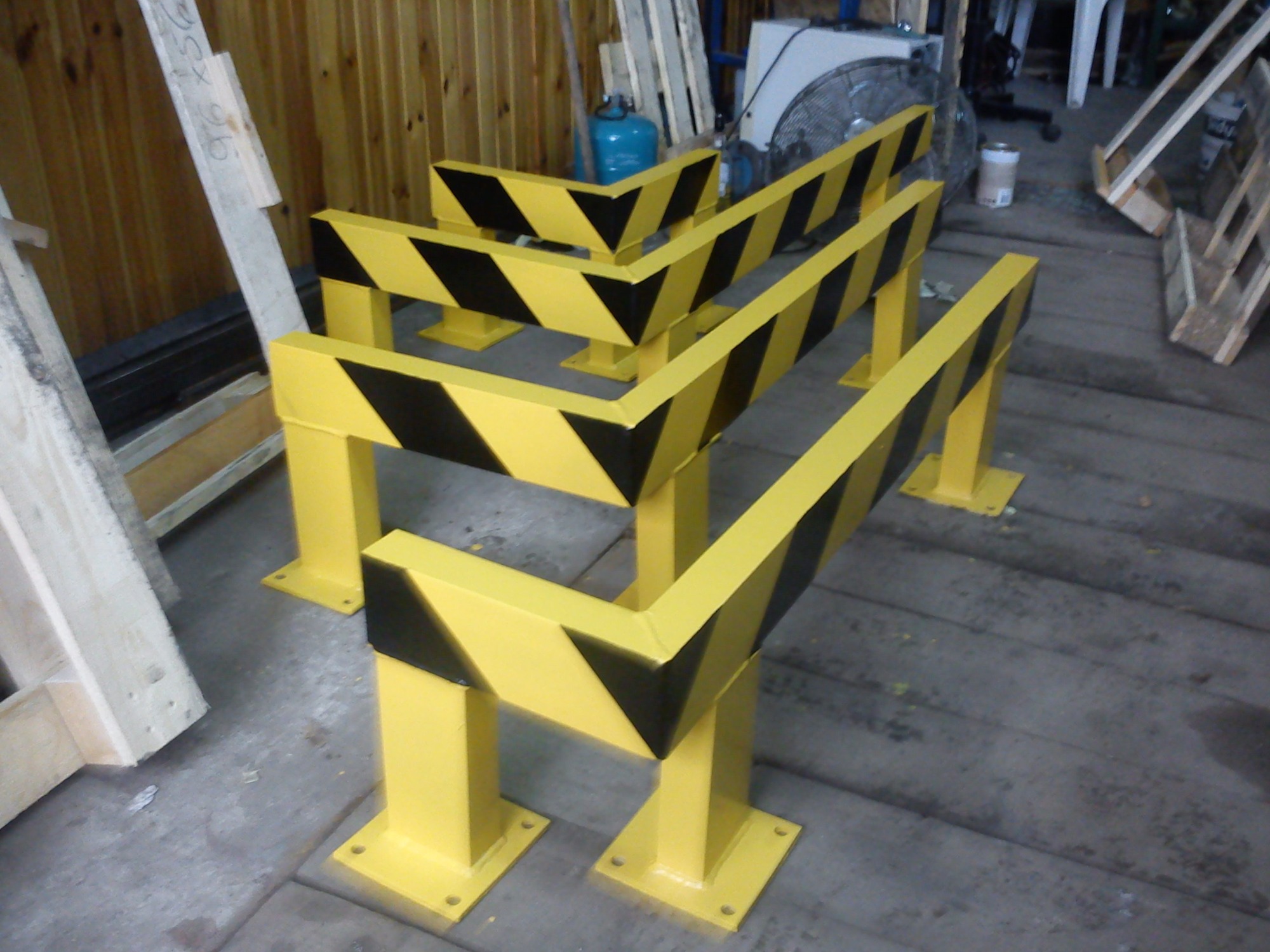 Żółte, stalowe bariery ochronne pomalowane w czarne pasy, ustawione na betonowej posadzce w hali produkcyjnej, w tle drewniane elementy konstrukcyjne i niebieska butla gazowa.