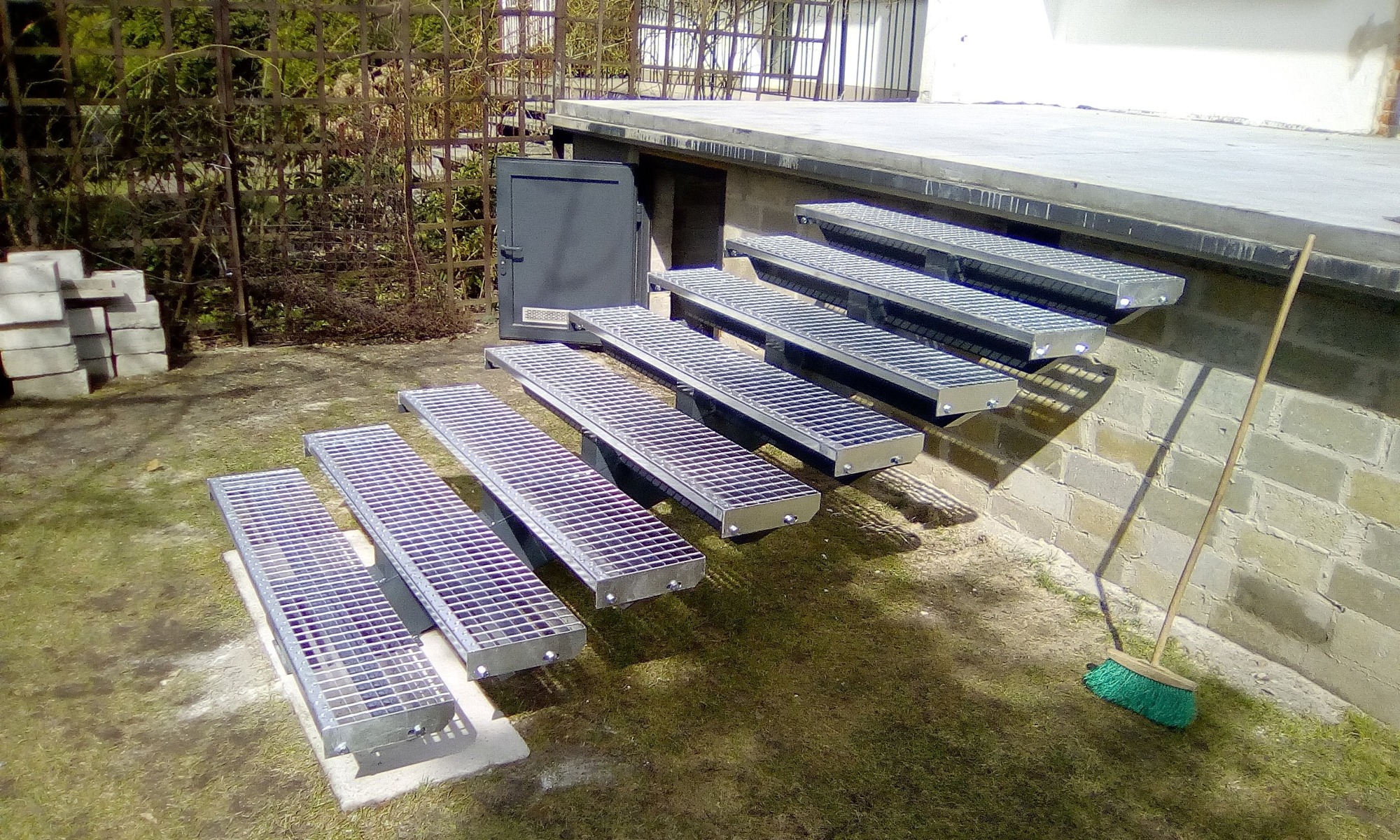 Zewnętrzne metalowe schody kratowe prowadzące do drzwi piwnicznych, widoczne świeże spawy na konstrukcji, obok ściany oparta miotła.