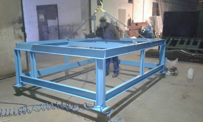 Niebieska, stalowa rama konstrukcyjna podnoszona przez suwnicę w hali produkcyjnej, widoczny pracownik w kombinezonie i elementy wyposażenia warsztatu w tle.