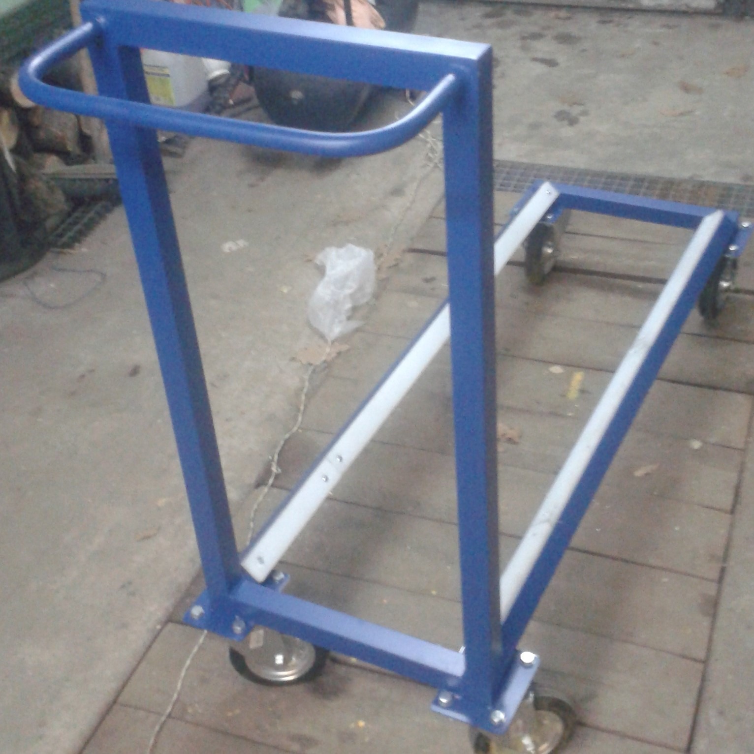 Wózek transportowy z niebieskich profili stalowych z białymi wzmocnieniami, na czterech kółkach, na tle betonowej posadzki.