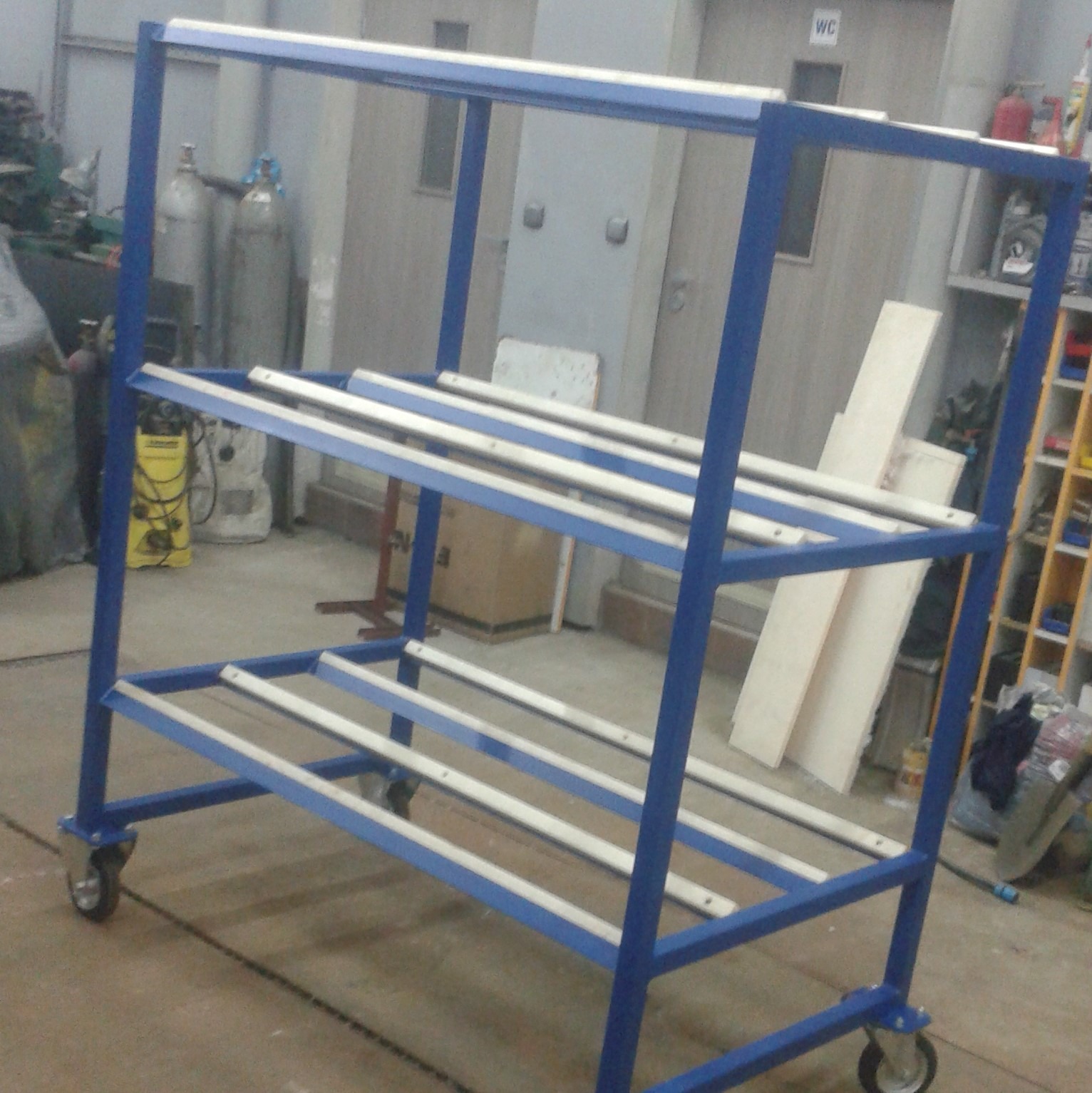 Niebieski, metalowy wózek transportowy na kółkach z dwoma poziomami, z białymi poprzeczkami, na tle warsztatu z butlami gazowymi i regałem.