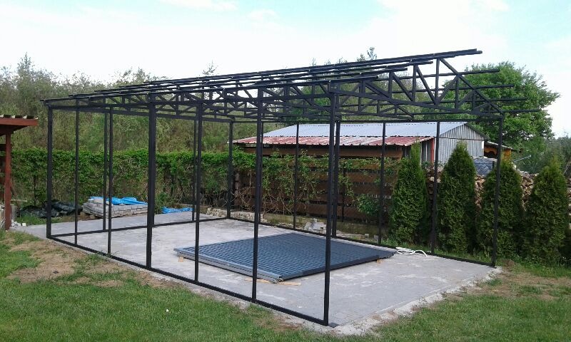 Stalowa konstrukcja z kratownicą dachową, prawdopodobnie zadaszenie tarasu lub wiaty, w trakcie montażu na betonowej wylewce, z widocznym fragmentem zwiniętej plandeki na podłożu i zieleni w tle.