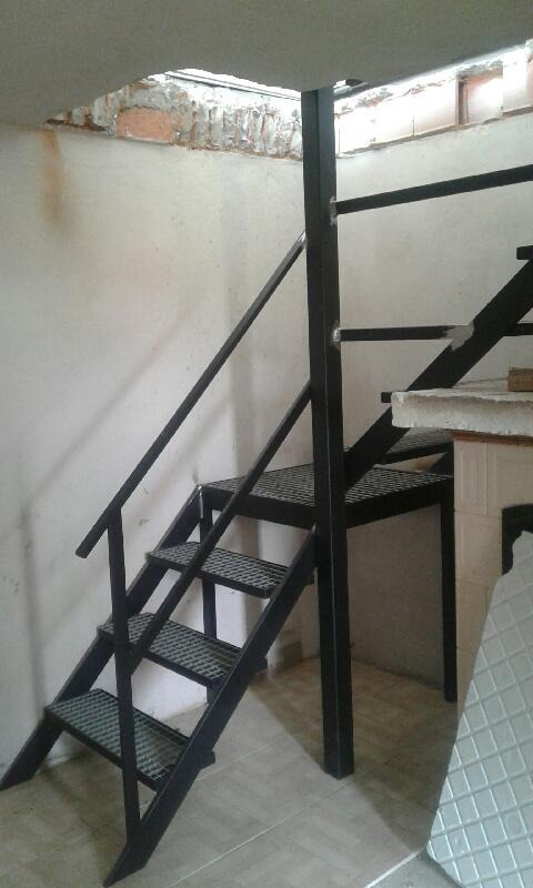 Metalowe schody z kratowymi stopniami, prowadzące do wyższej kondygnacji w surowym wnętrzu z otynkowaną ścianą i widocznymi fragmentami cegieł.