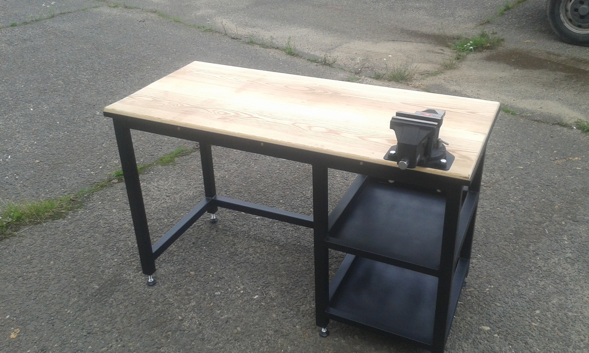 Stół warsztatowy z drewnianym blatem i czarną, metalową konstrukcją, wyposażony w imadło, na zewnątrz.