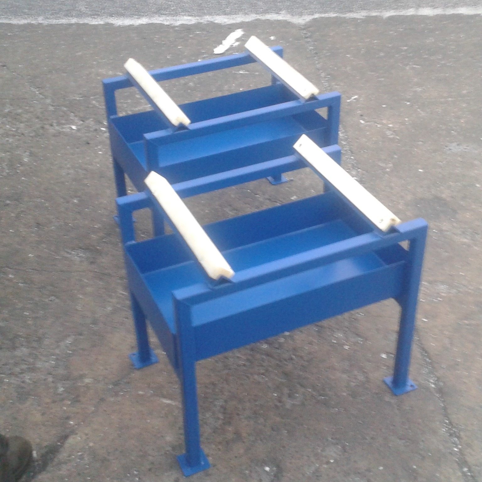 Dwa niebieskie, spawane stojaki z metalu, z białymi elementami zabezpieczającymi na górze, stojące na betonowej nawierzchni, widoczne z perspektywy lekko z góry.