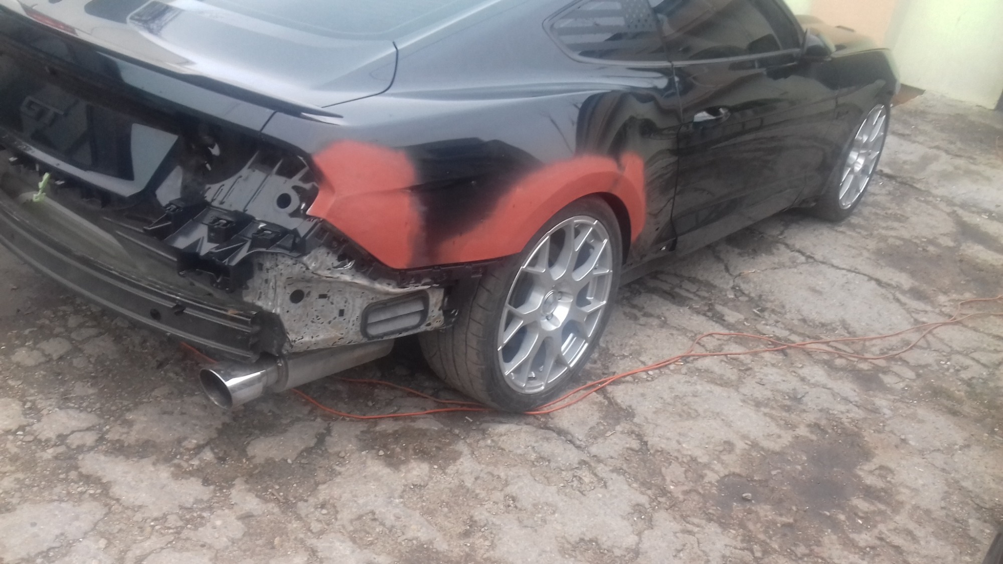 Czarny samochód w trakcie naprawy blacharskiej, z widocznymi obszarami pokrytymi podkładem lakierniczym, odsłoniętym tylnym zderzakiem i fragmentem układu wydechowego, na betonowym podłożu.