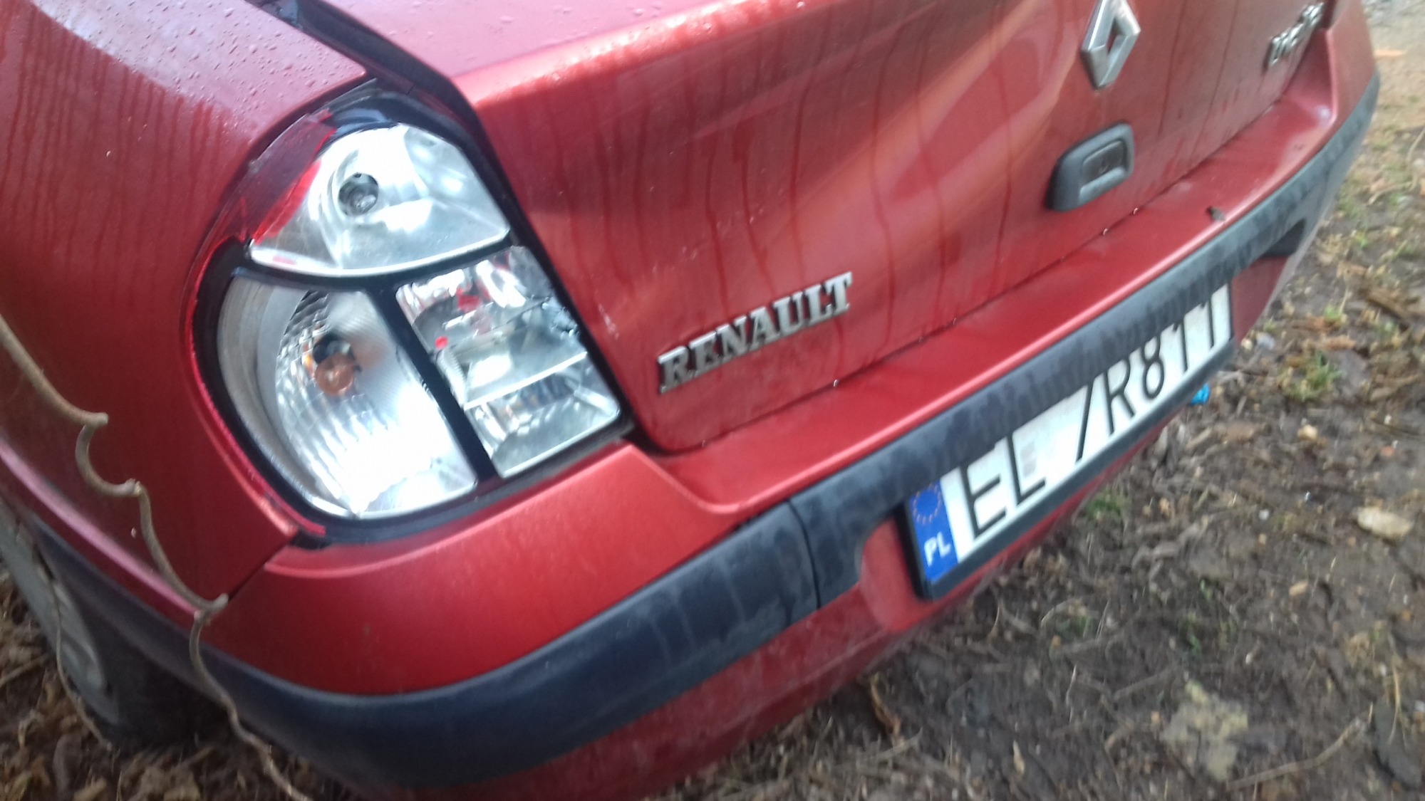 Czerwony Renault Clio z uszkodzonym tylnym światłem, widoczne ślady błota na karoserii i ziemi wokół pojazdu.
