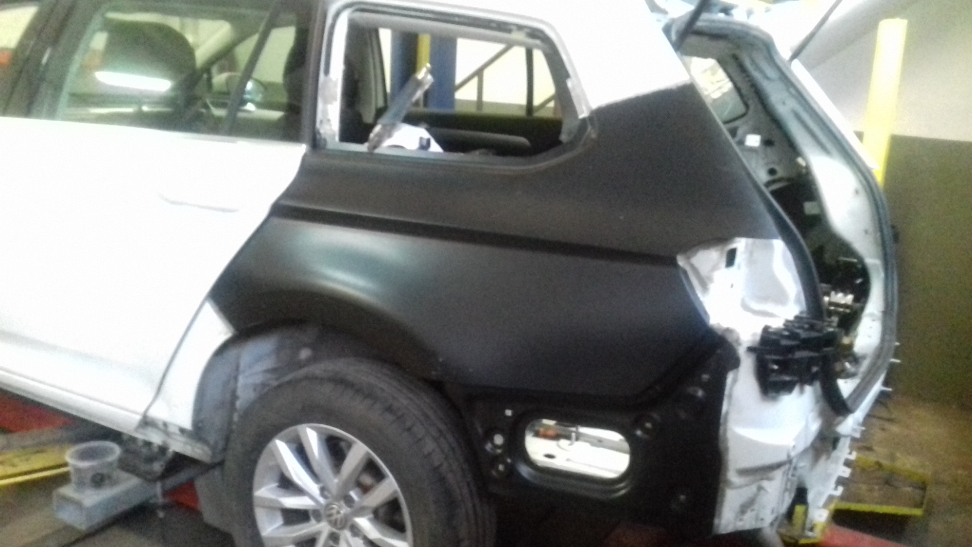 Biały samochód kombi w trakcie naprawy blacharskiej: brak tylnej klapy i fragmentu karoserii, widoczne elementy konstrukcyjne, czarny element karoserii w trakcie montażu, koło z felgą aluminiową.