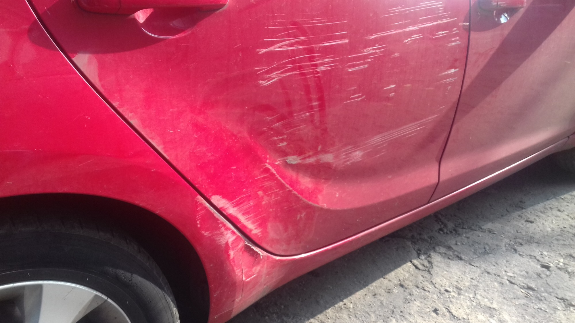 Czerwony samochód z licznymi zarysowaniami na tylnych drzwiach i błotniku, widoczne uszkodzenia lakieru.