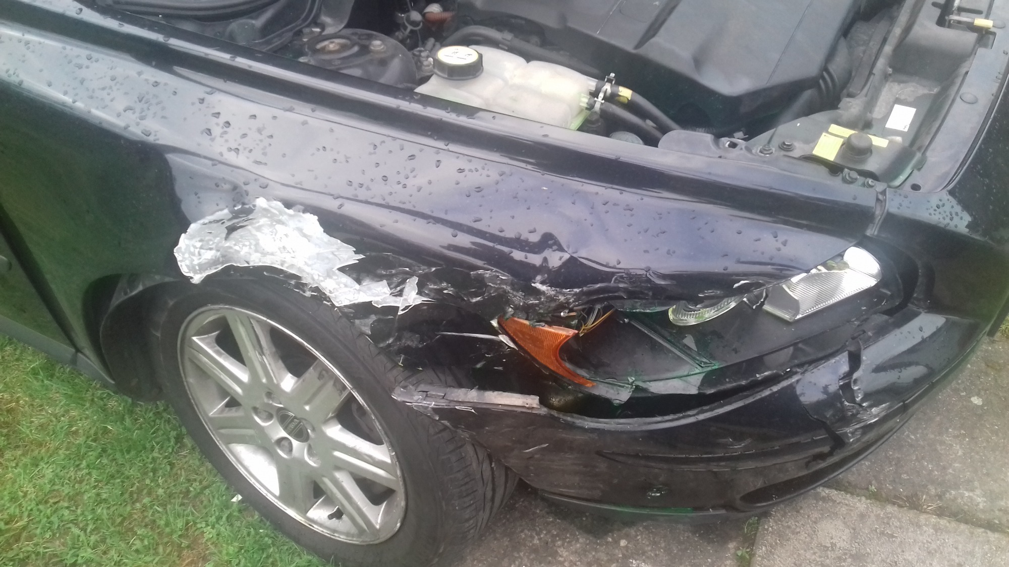 Uszkodzony przód czarnego samochodu po kolizji, widoczne zgniecione blachy, pęknięty reflektor i odklejony lakier na błotniku, w tle widoczny silnik.