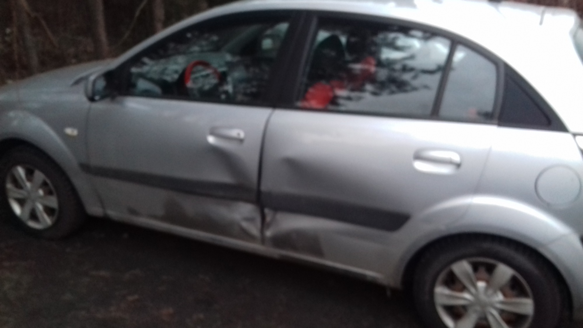 Srebrny samochód z wyraźnymi wgnieceniami na drzwiach i progu, widoczne czerwone akcenty wewnątrz pojazdu, zaparkowany na tle drzew.