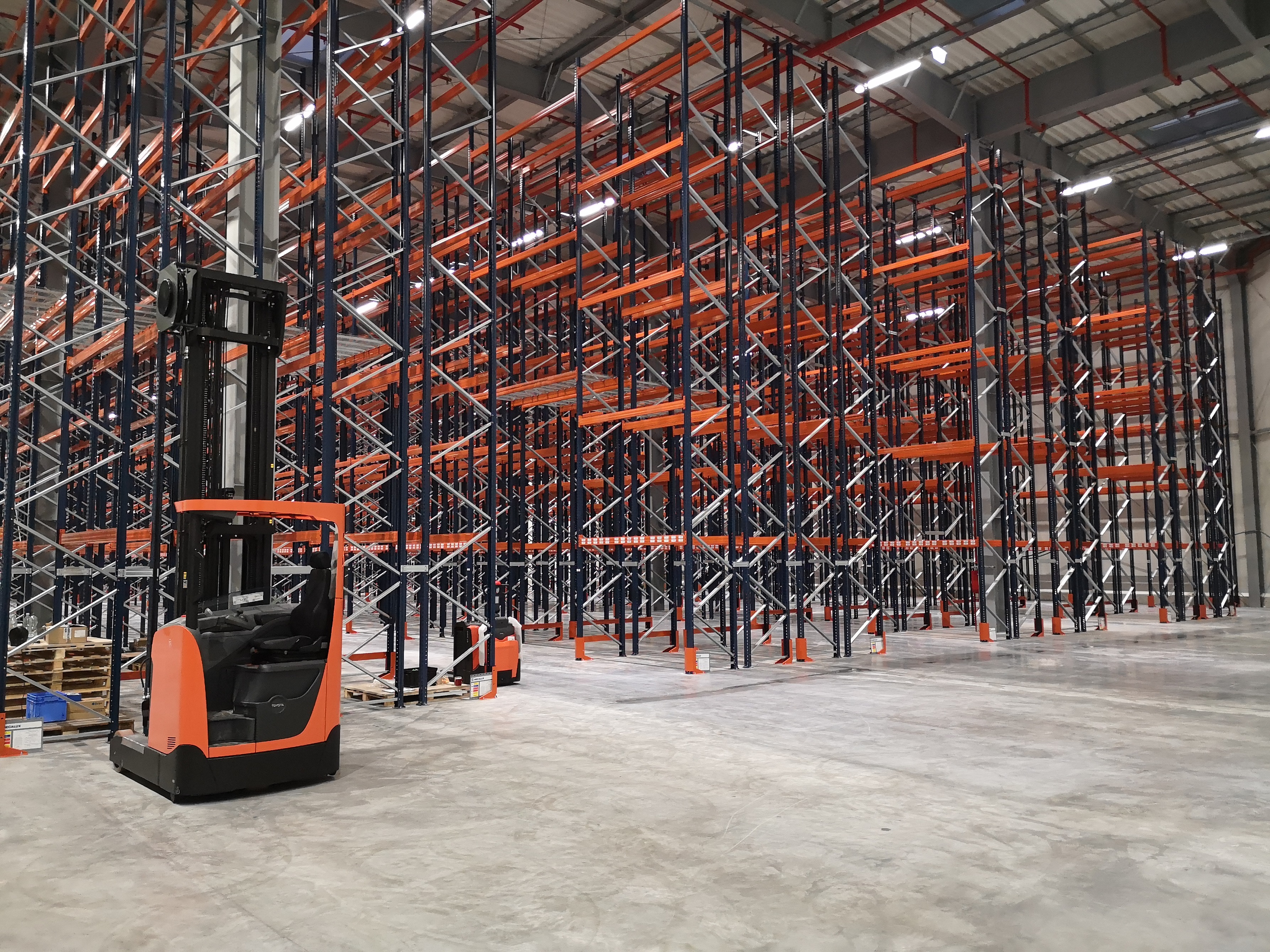 Wnętrze magazynu z wysokimi regałami metalowymi w kolorach czarnym i pomarańczowym oraz wózkiem widłowym Toyota w kolorze pomarańczowo-czarnym na pierwszym planie.
